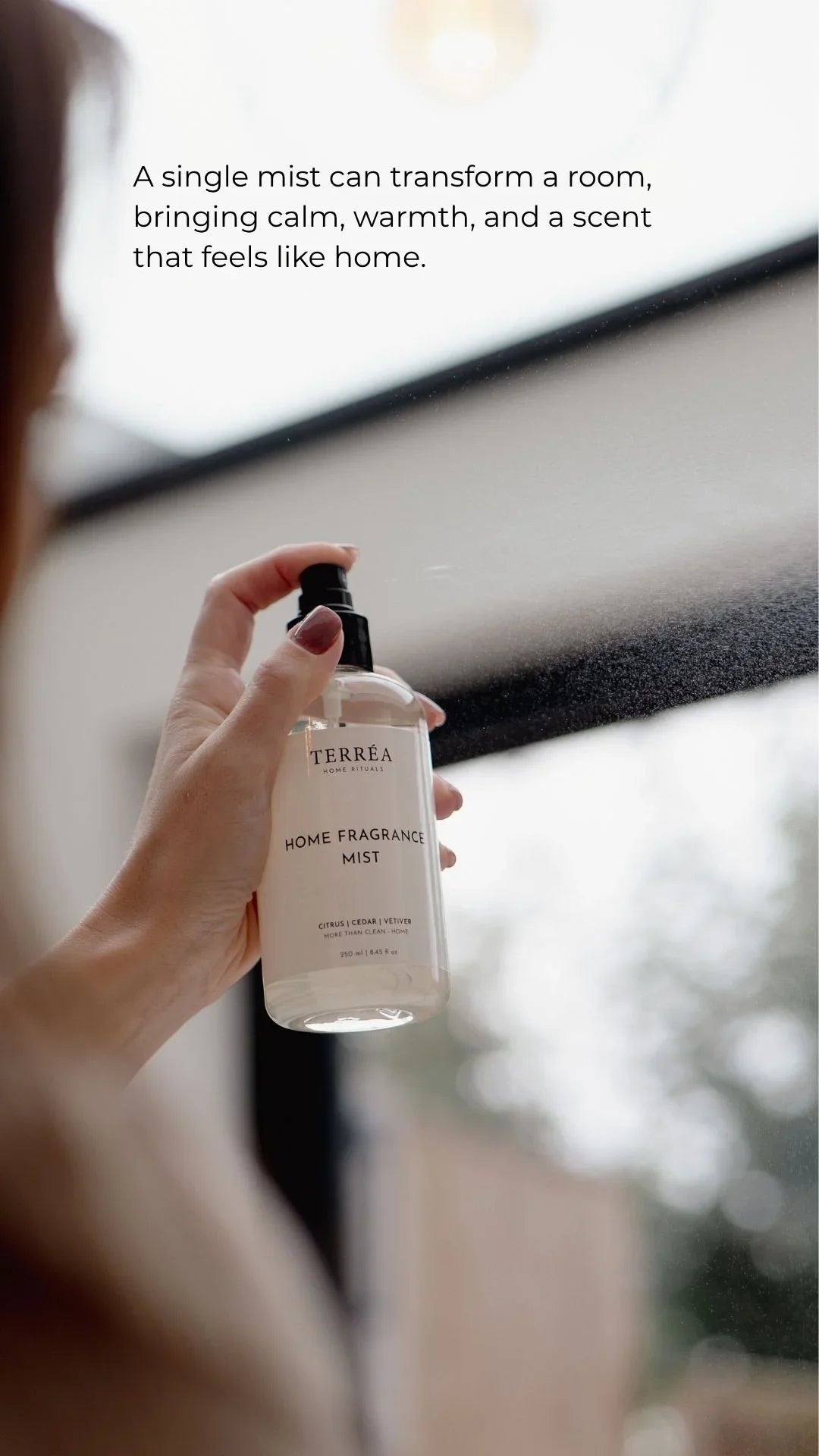 Hand holding TERRÉA Home Fragrance Mist spray bottle, misting for calm, warm, homey scent near window