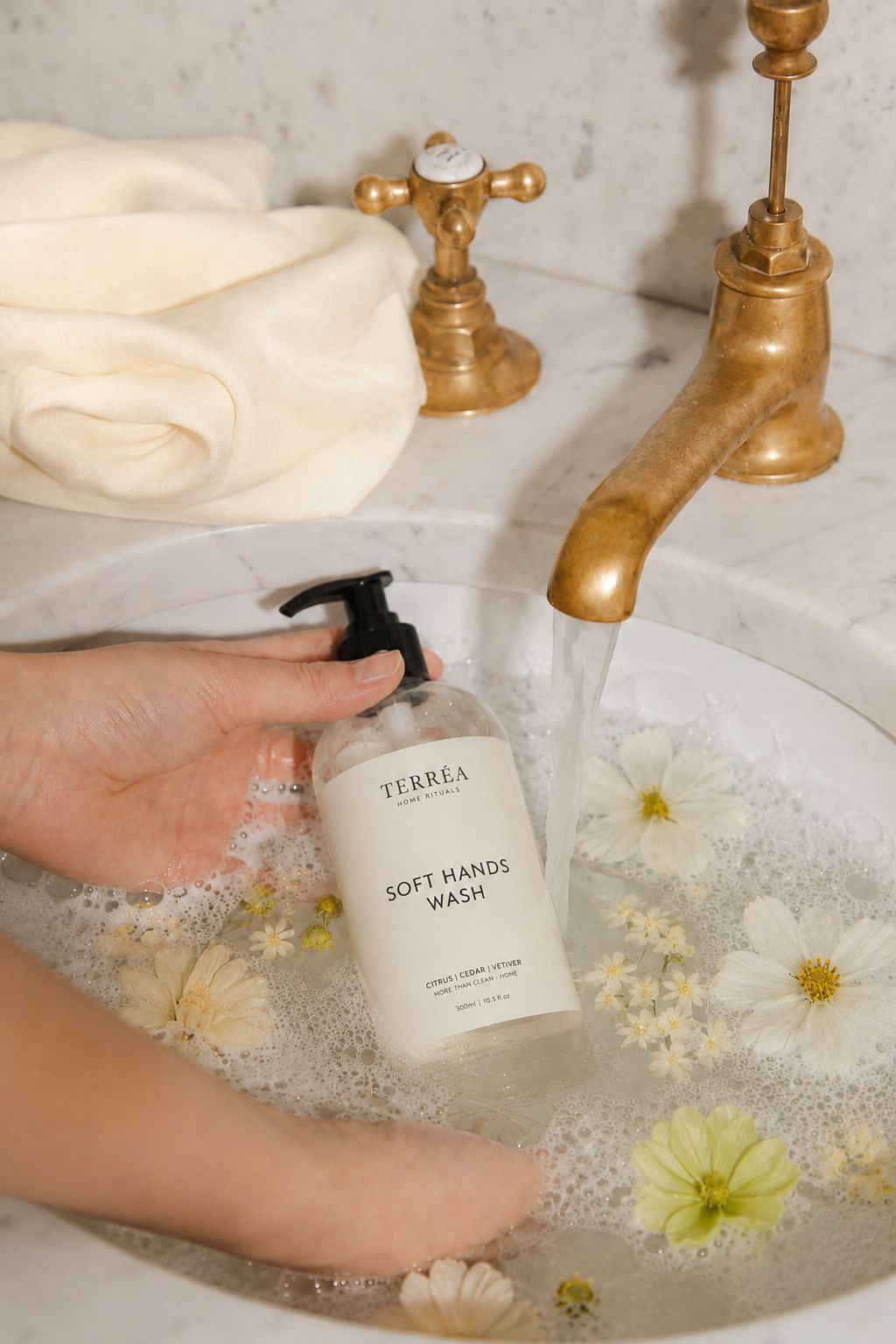 Terréa Soft Hands Wash bottle held under running water in a marble sink filled with soft foam and floating white flowers.