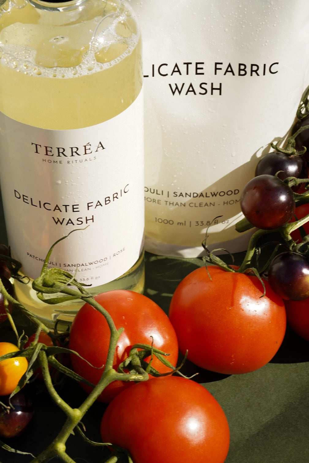 Terréa Delicate Fabric Wash bottle and refill pouch styled with ripe tomatoes and dark berries against an olive-green backdrop, with warm sunlight highlighting fresh droplets and glossy surfaces.