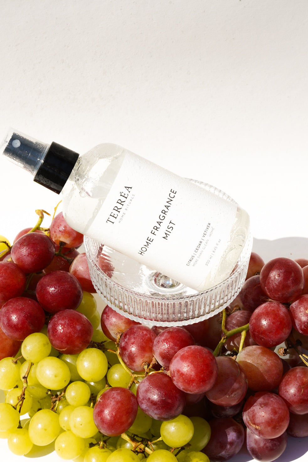 Terréa Home Fragrance Mist in Citrus, Cedar and Vetiver resting on a glass dish among red and green grapes, with bright sunlight highlighting fresh droplets on the bottle.