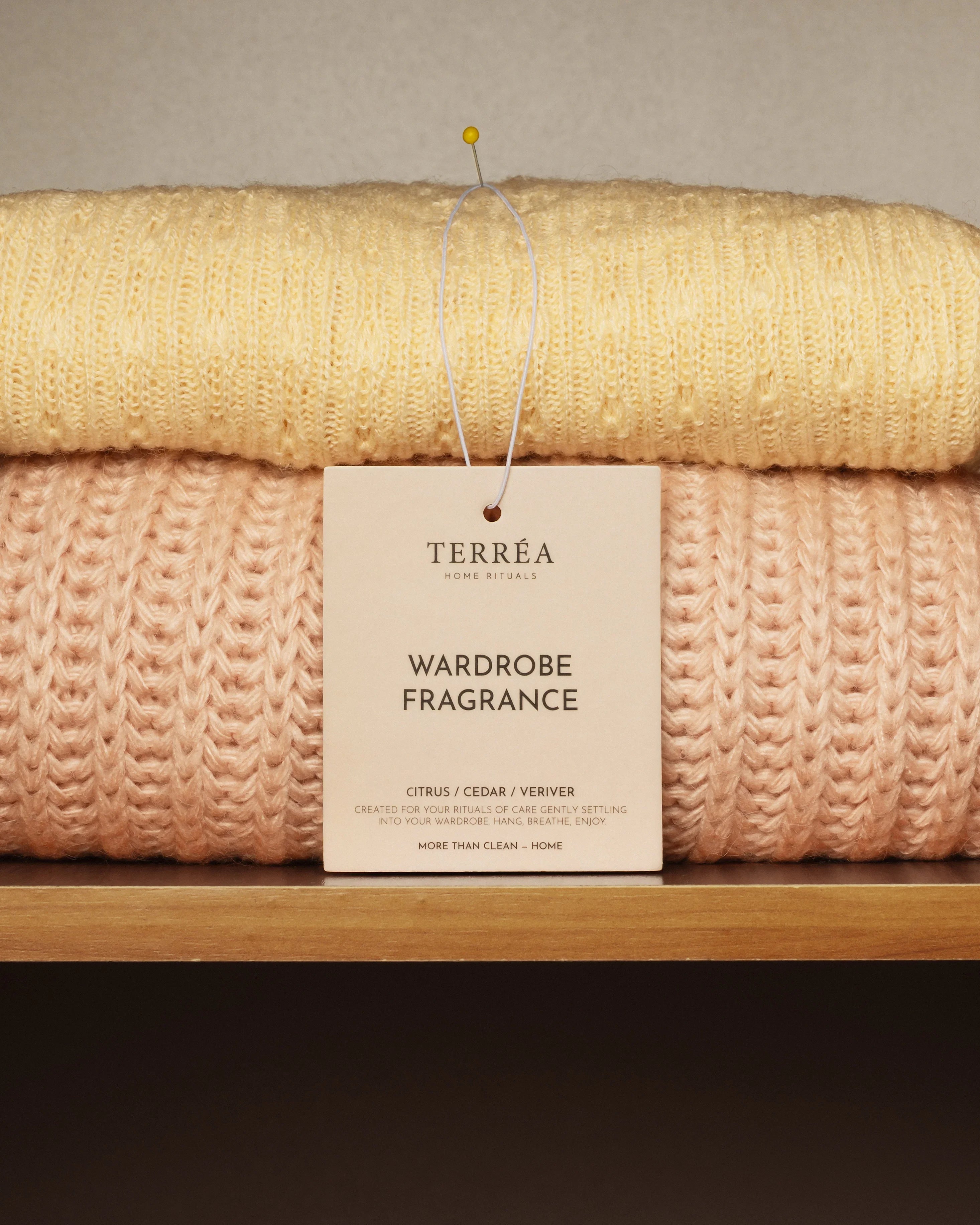 Folded jumpers (yellow, peach) on a shelf with Terréa Wardrobe Fragrance, Citrus/Cedar/Vetiver scent.