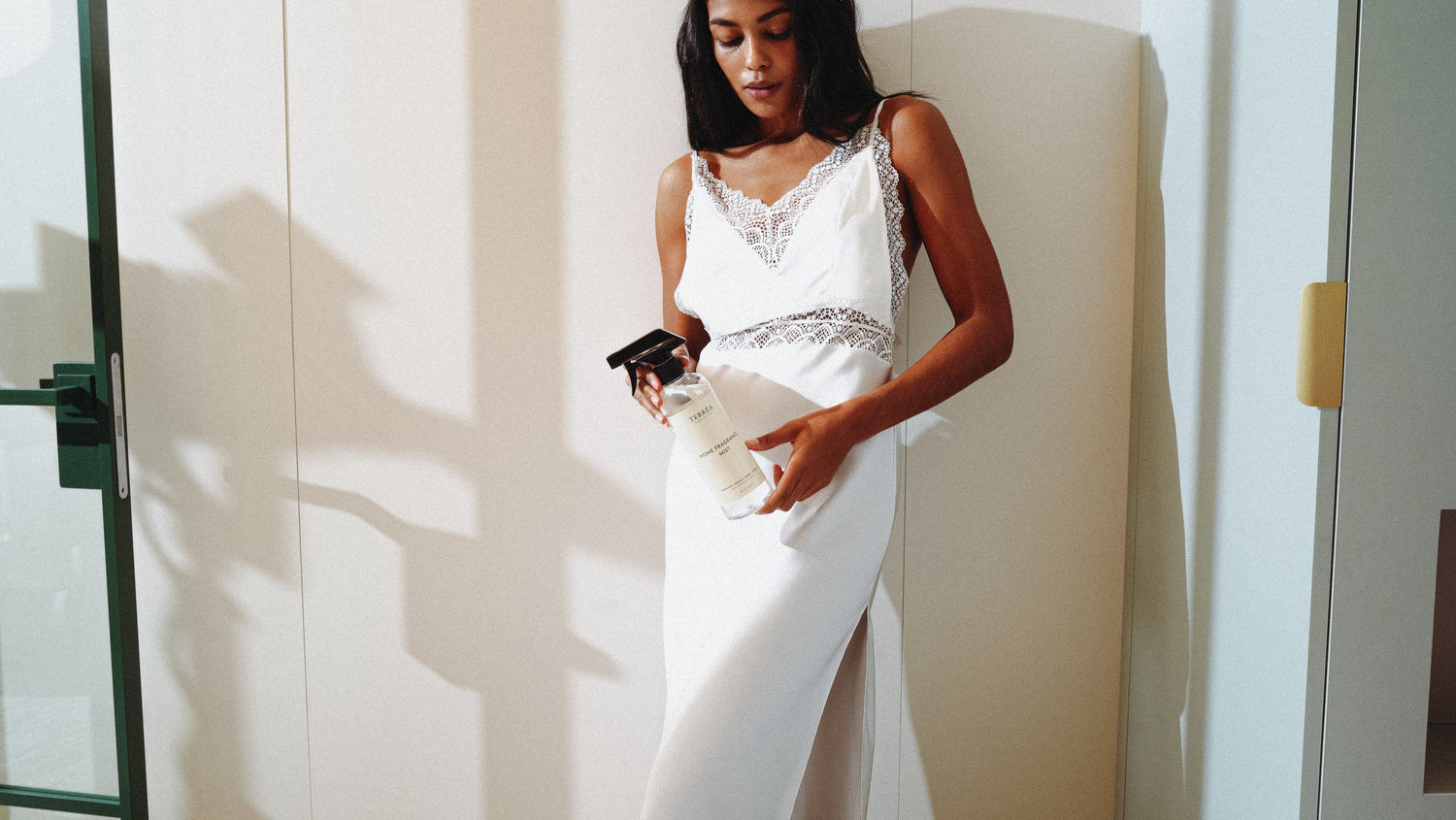 Woman holding eco-friendly home fragrance spray in white lace dress, natural light setting