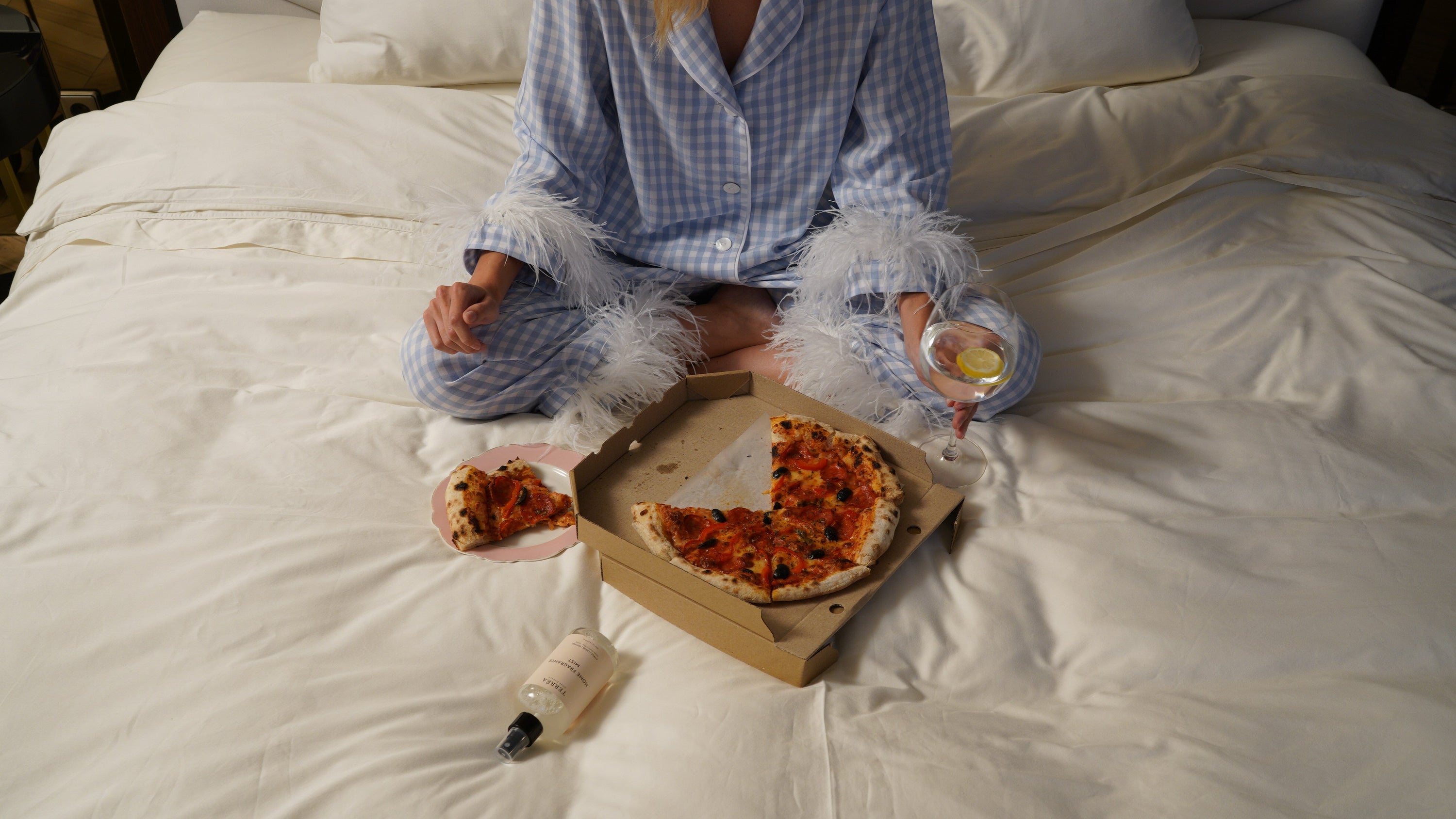 Woman in checkered pajamas enjoying pizza and drink on cozy bed with home fragrance mist