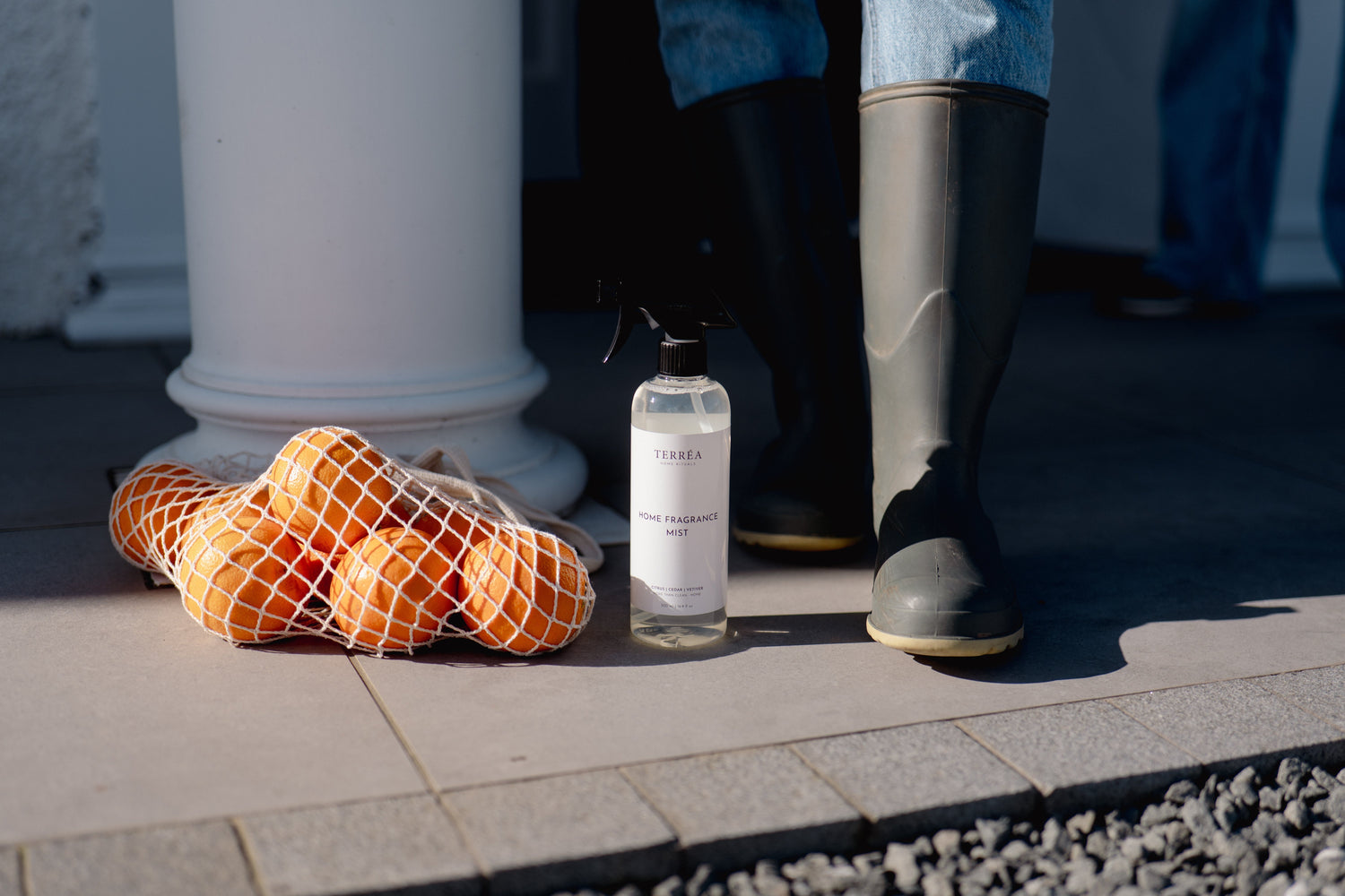 Home fragrance mist bottle beside oranges in net bag, near boots on patio