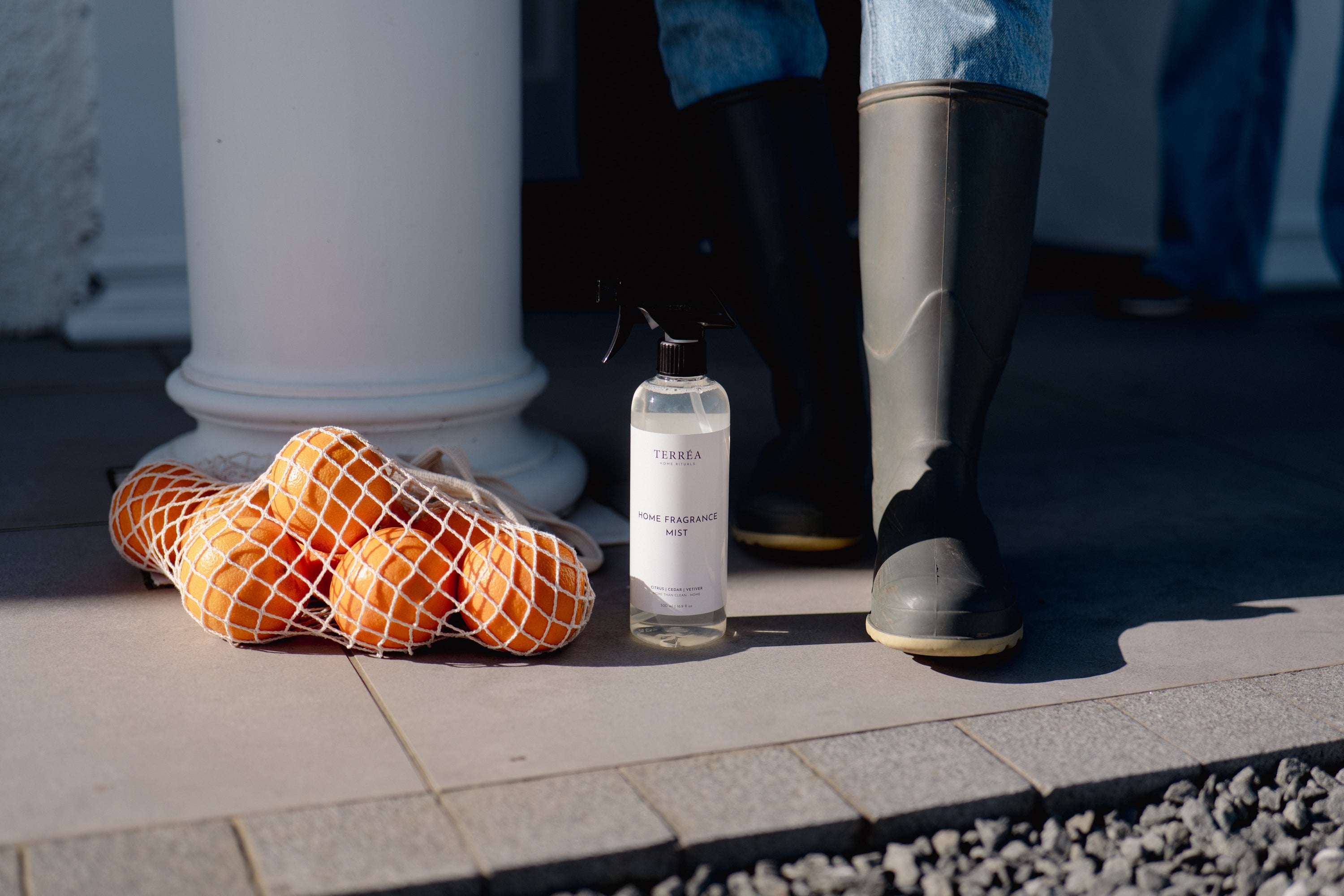 Home fragrance mist bottle beside oranges in net bag, near boots on patio