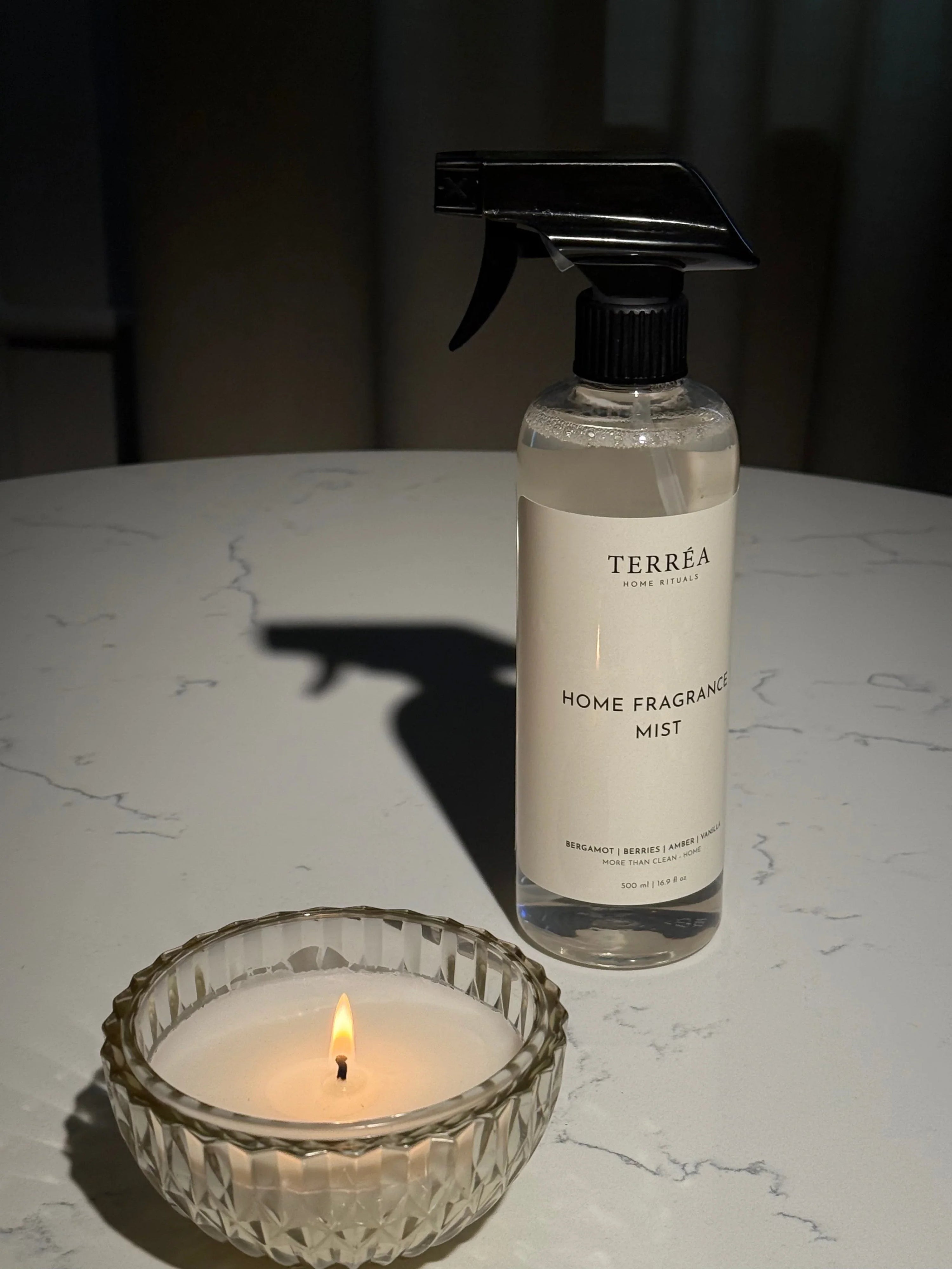 Home fragrance mist spray bottle beside lit candle on marble surface, eco-conscious home ritual