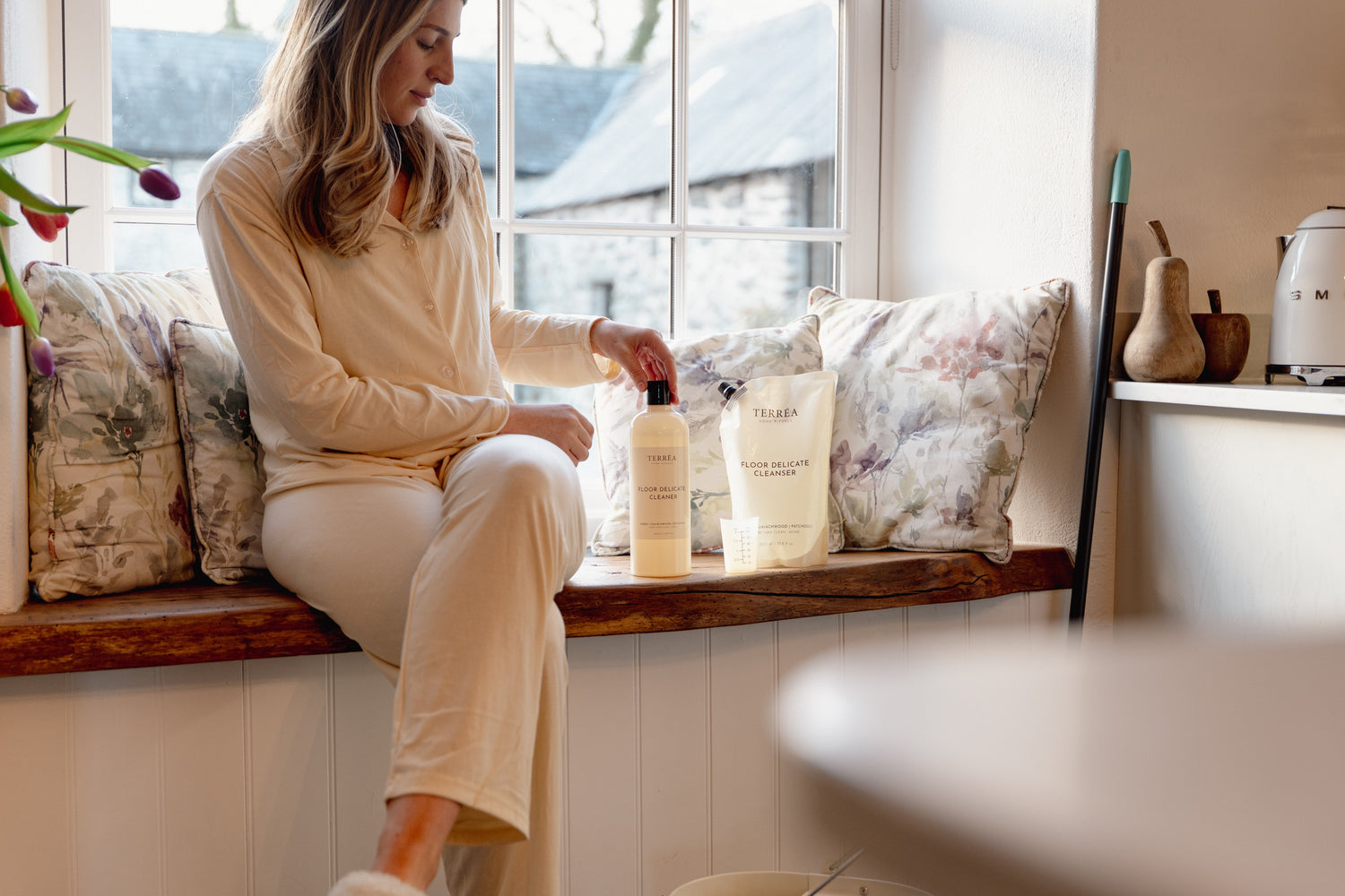 Woman holding eco-friendly floor cleaner bottle near window with floral pillows and home decor