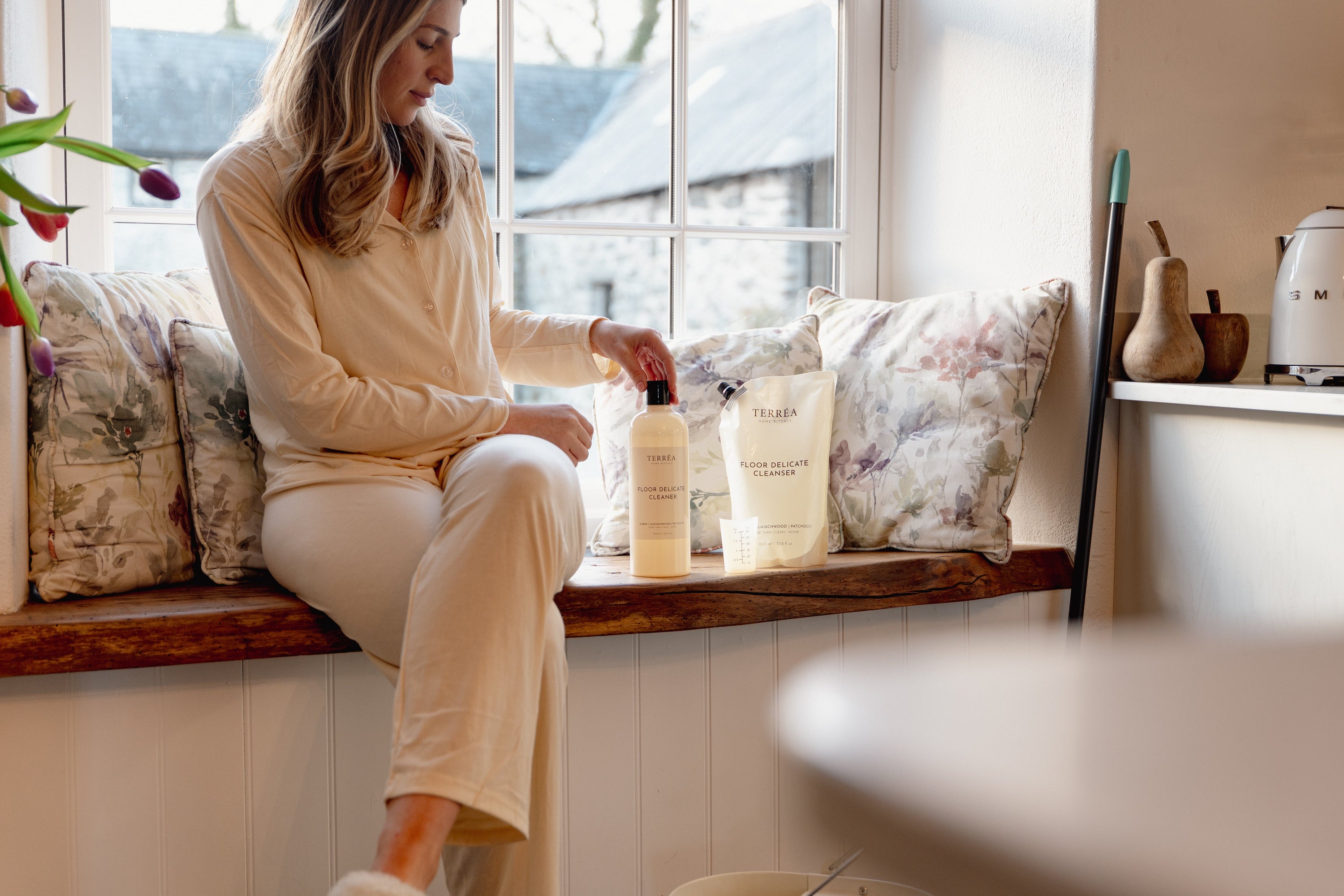 Woman holding eco-friendly floor cleaner bottle near window with floral pillows and home decor