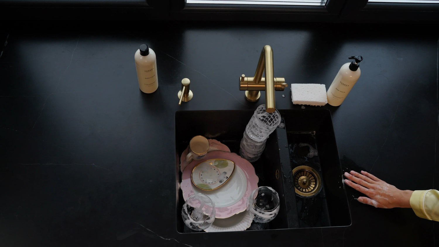 Overhead view of hand dishwashing at a black sink with a brass tap and Terréa Dish Calm bottles on the worktop
