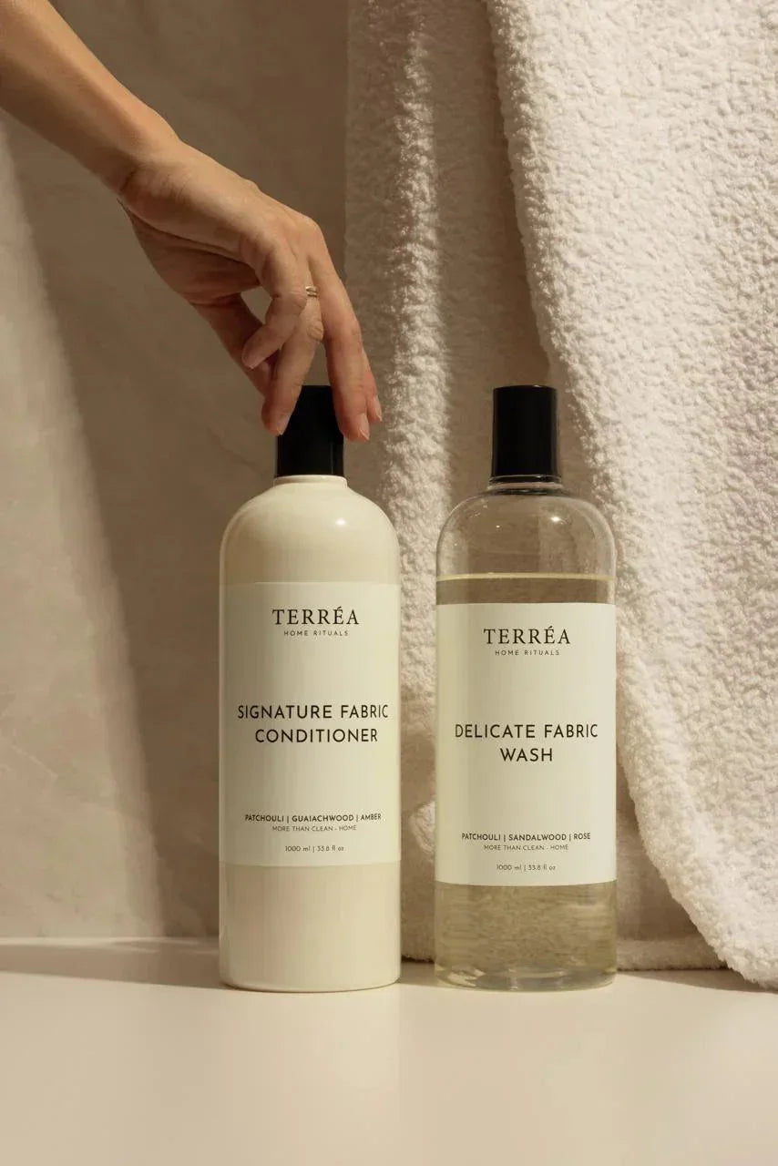 Signature Fabric Conditioner and Delicate Fabric Wash bottles with hand touching cap, soft towel background