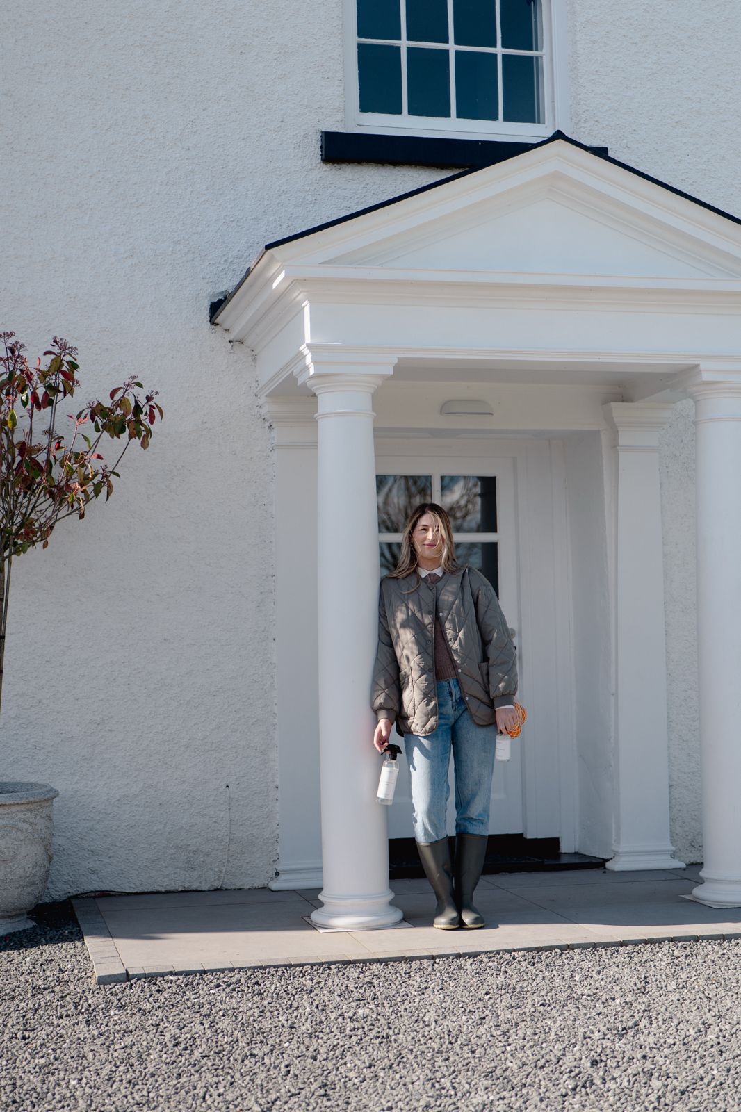 Woman at a white porch holding Terréa Clean Home Surfaces 500 ml spray and coiled rope, gravel driveway in sunlight