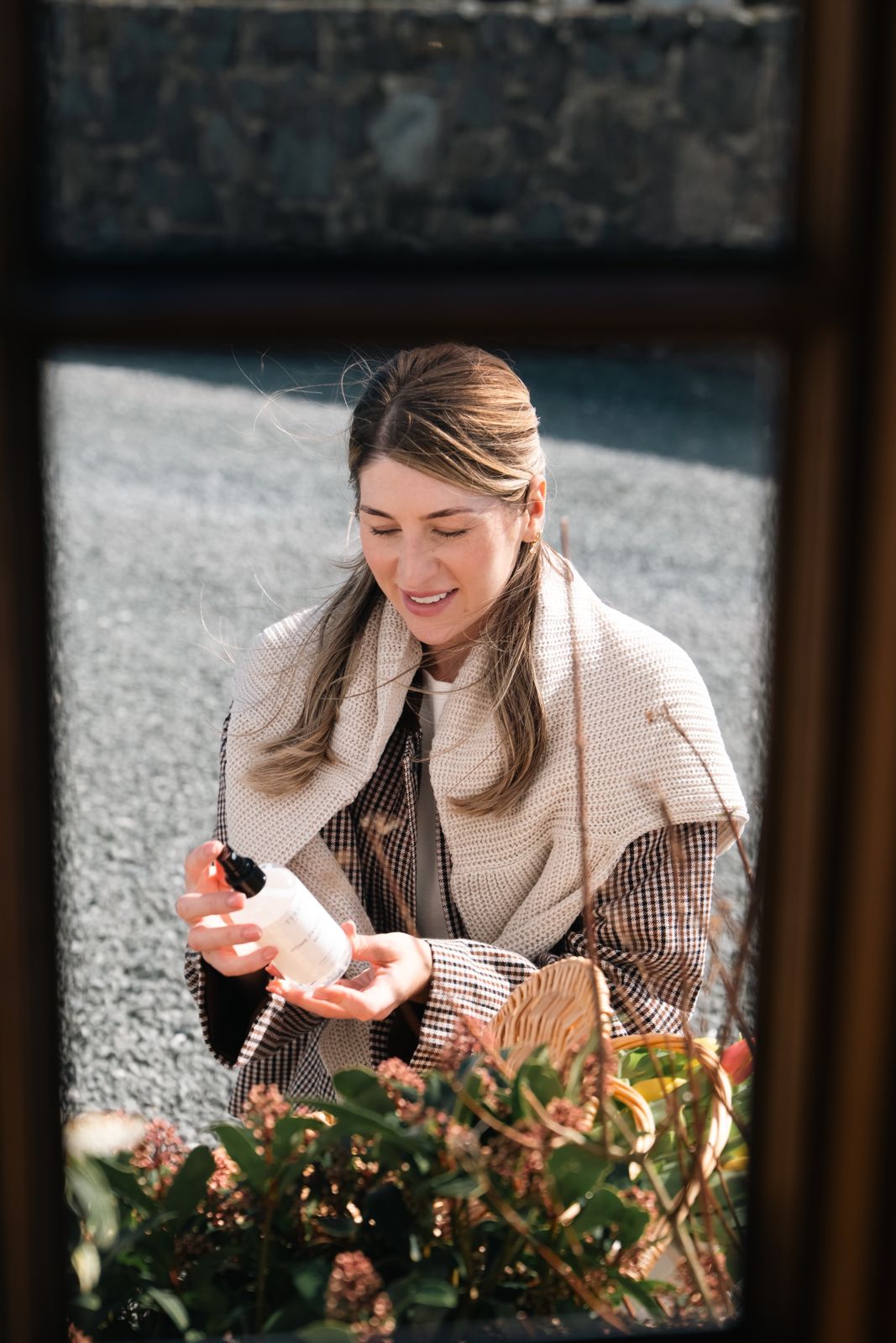 Woman holding Terréa Home Fragrance Mist 250 ml outdoors beside a basket of flowers, sunlit gravel in background