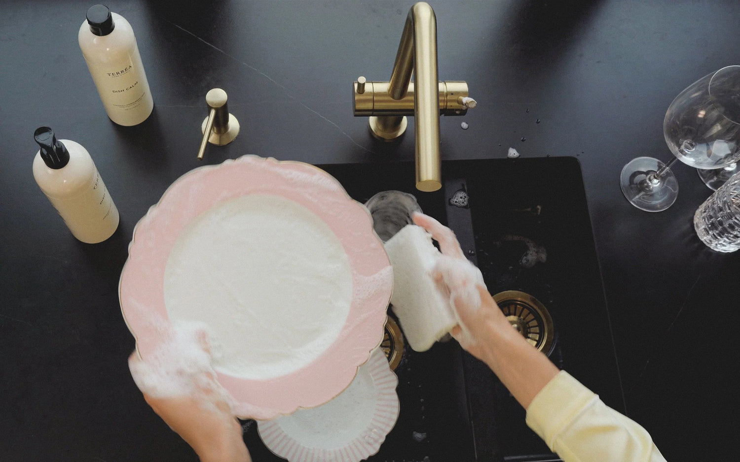 Hands washing pink scalloped plate with eco dish soap at black sink, gold faucet, sustainable kitchen care