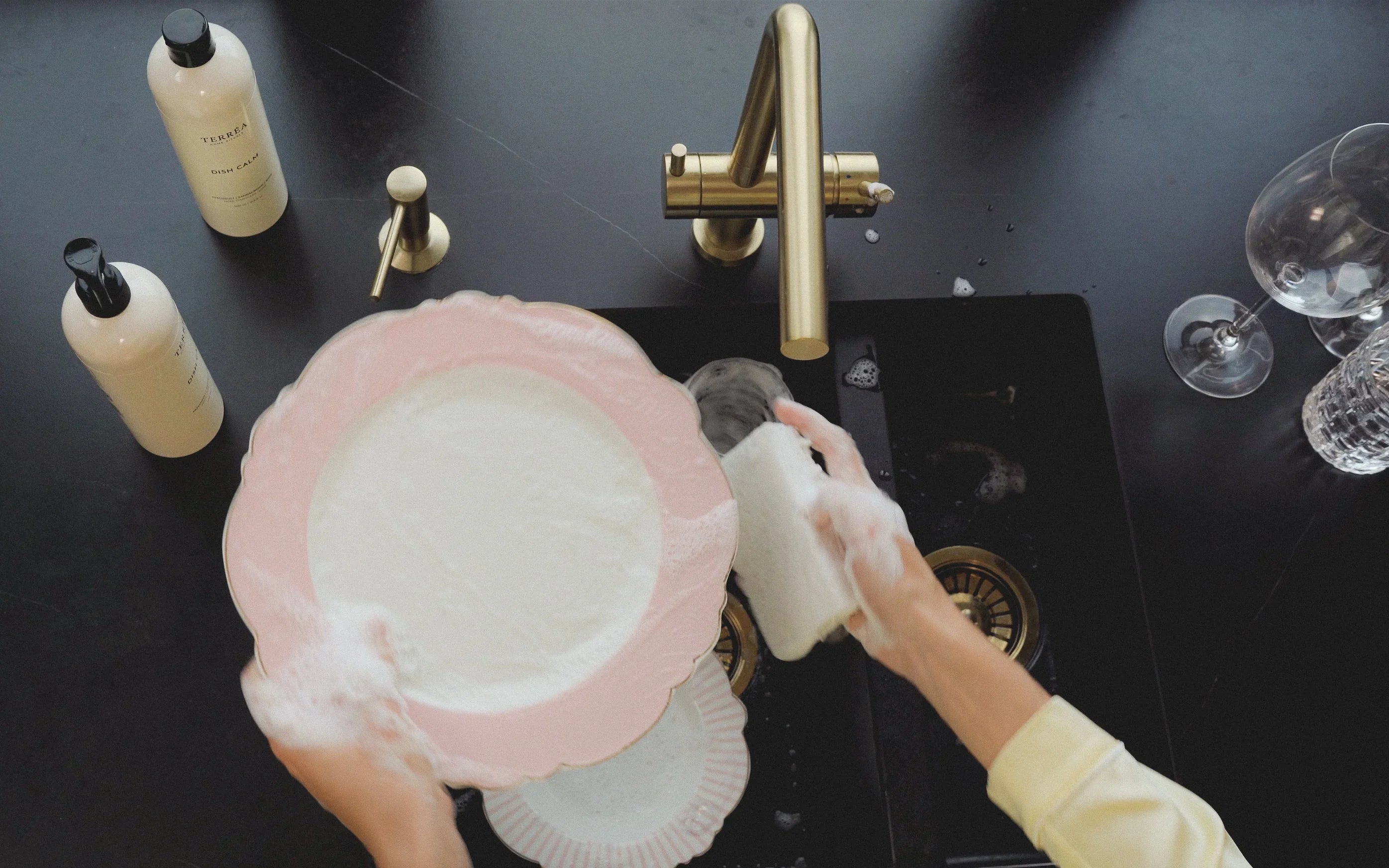 Hands washing pink scalloped plate with eco dish soap at black sink, gold faucet, sustainable kitchen care