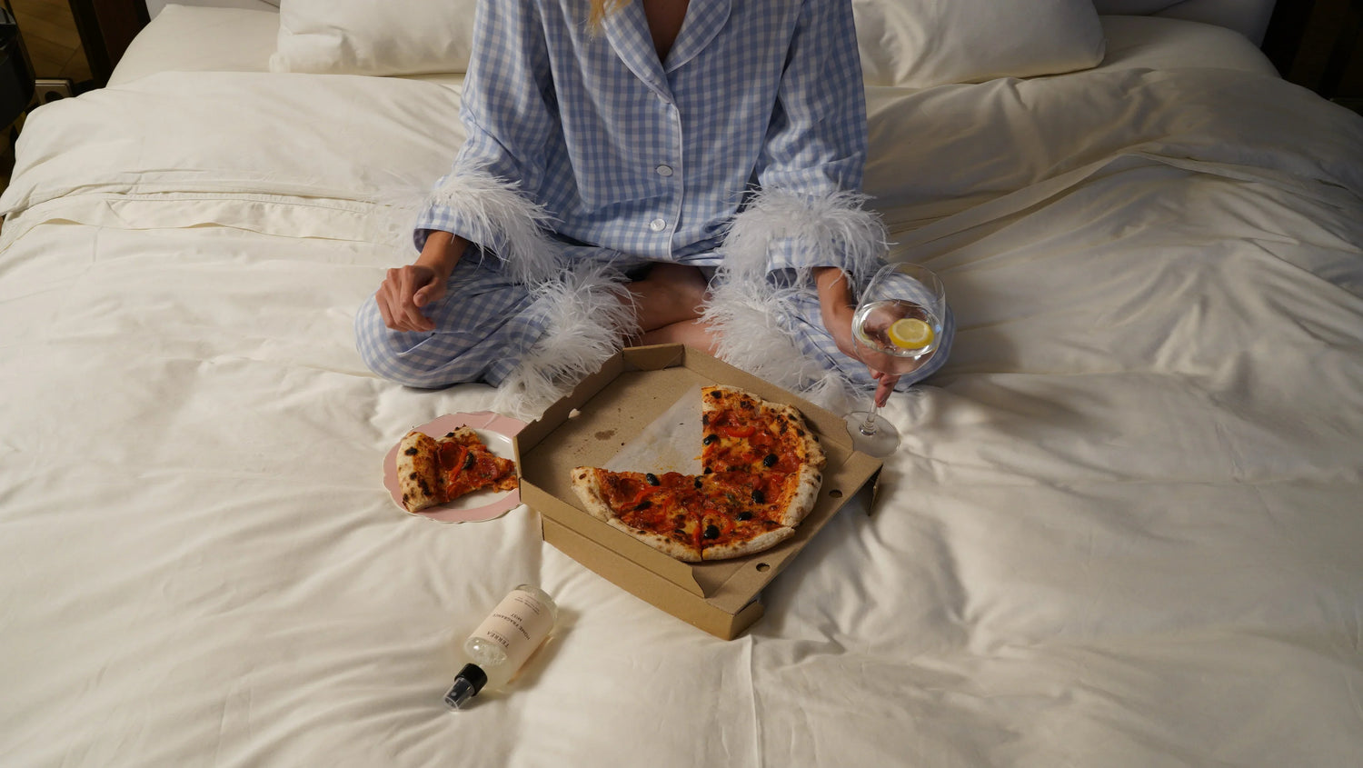 Woman on bed in a bedroom, eating pizza and holding a Terréa wardrobe freshener; glass of water nearby