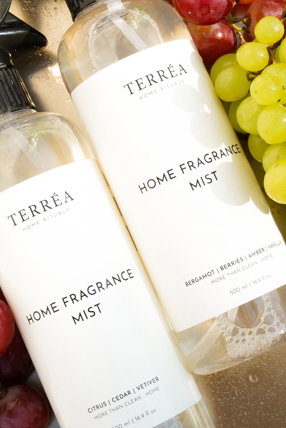 A close-up of two Terréa Home Fragrance Mist bottles nestled among red and green grapes, with golden light catching the liquid and bubbles.