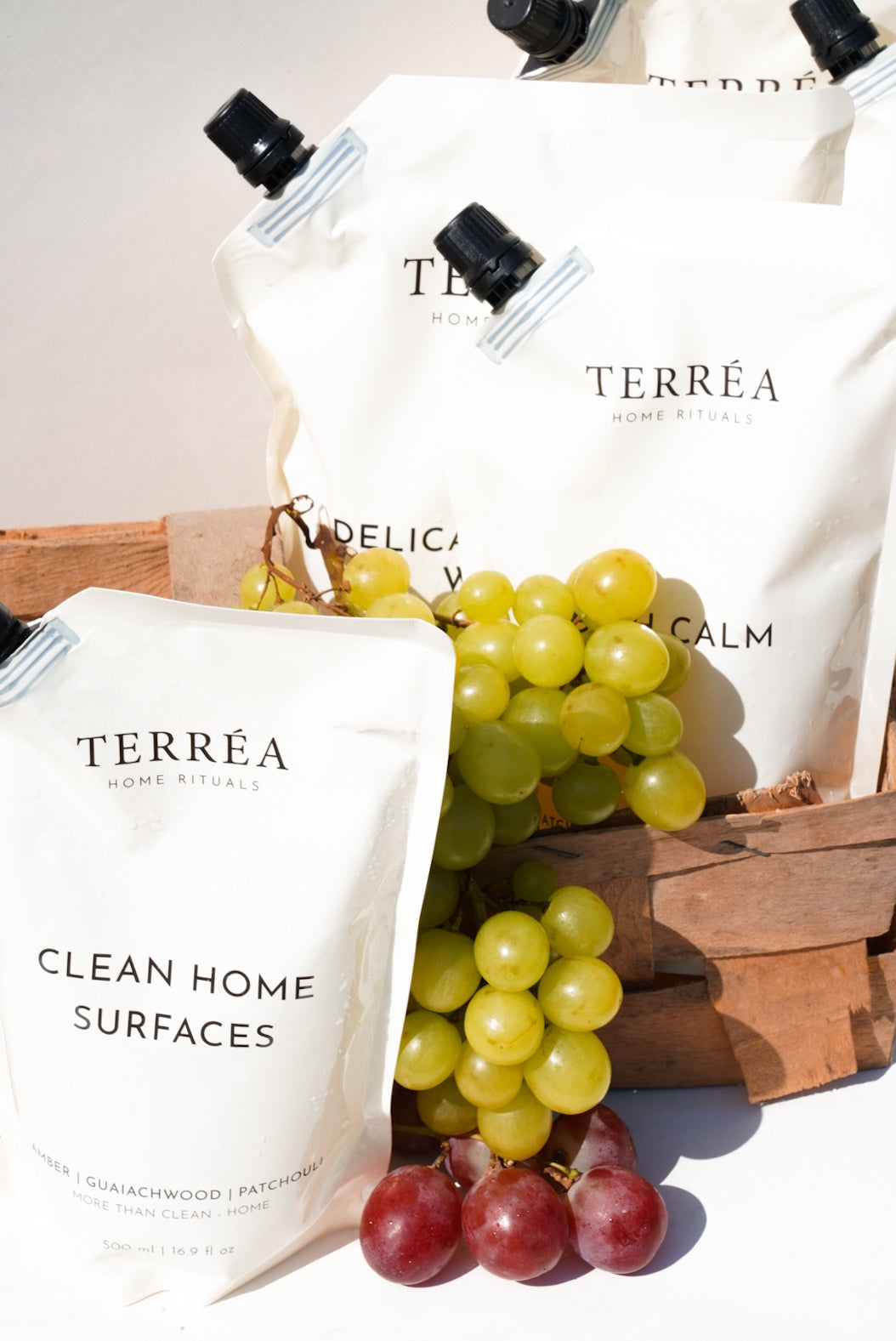 Terréa refill pouches for Clean Home Surfaces, Dish Calm and Delicate Fabric Wash styled in a wooden crate with green and red grapes in bright sunlight.
