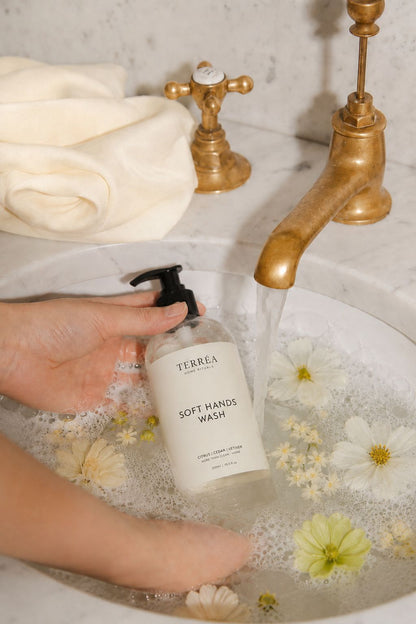 Terréa Soft Hands Wash bottle held under running water in a marble sink filled with soft foam and floating white flowers.