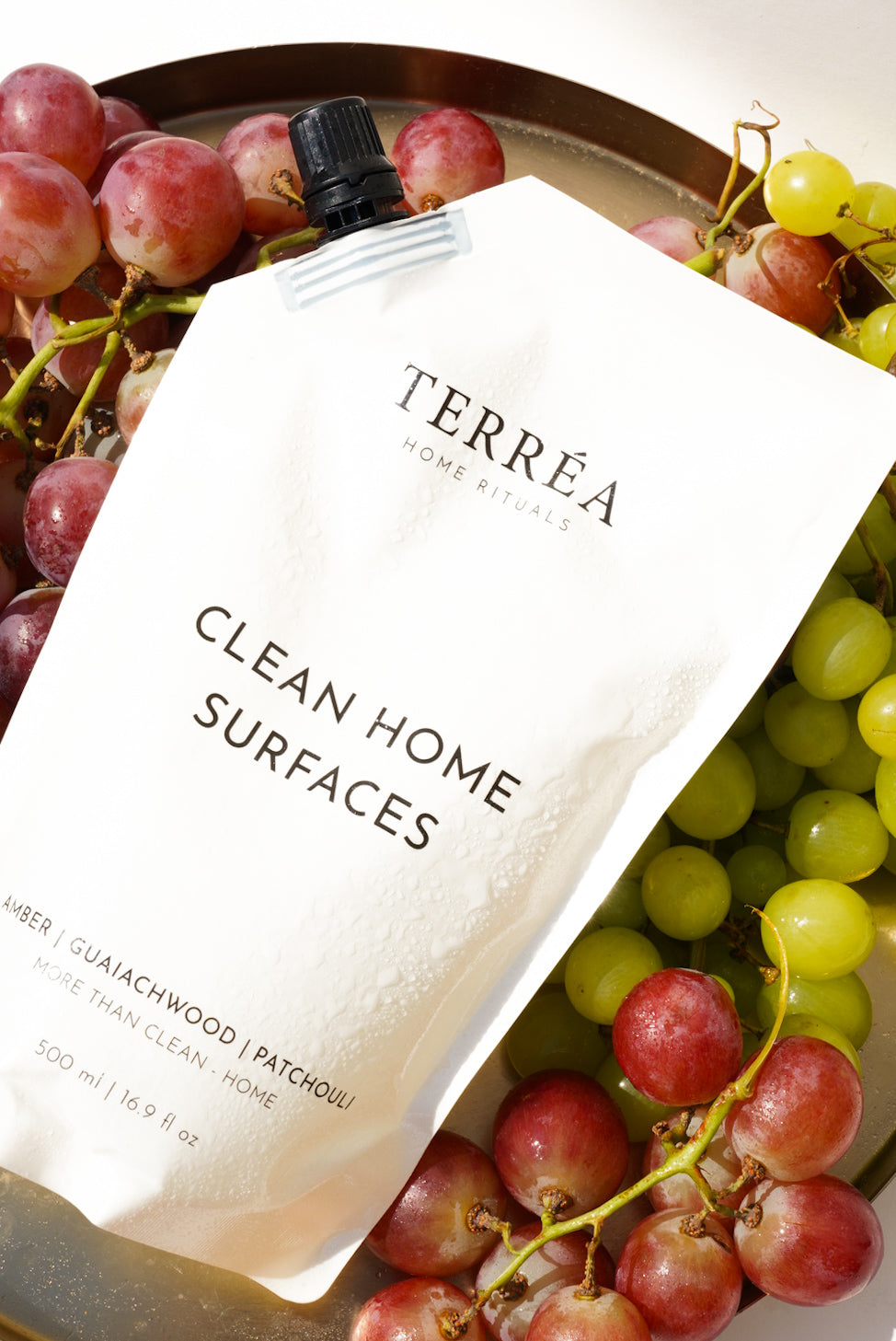 Terréa Clean Home Surfaces refill pouch styled on a golden tray with red and green grapes, with fresh droplets catching the warm sunlight.