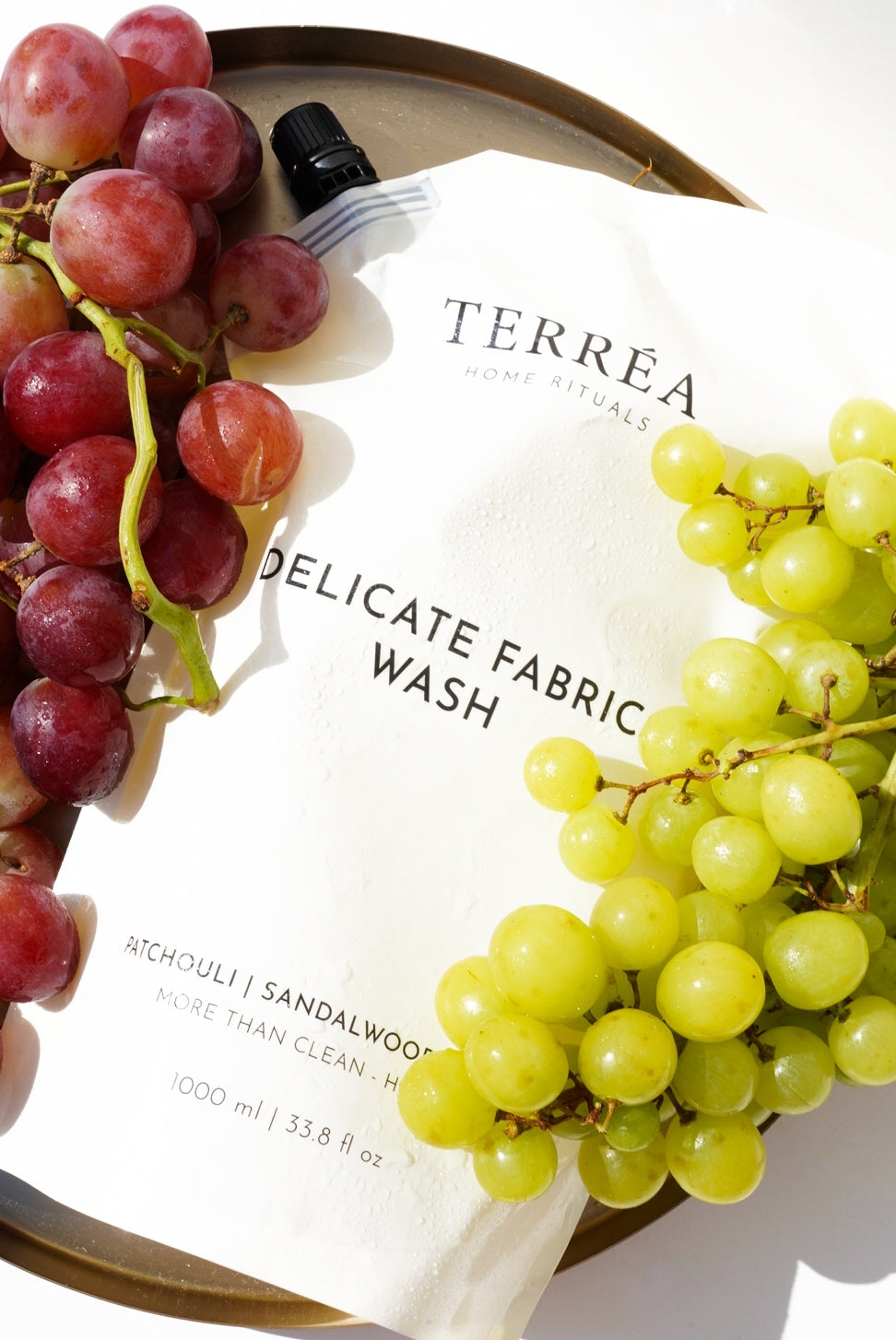Terréa Delicate Fabric Wash refill pouch styled on a golden tray with red and green grapes, lit by bright sunlight and soft shadows.