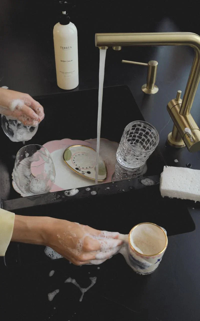 Person washing a ceramic cup in a black sink with a gold faucet, using a bottle of soap labeled 'Terras'.