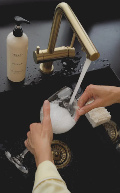 Person washing a white object under running water from a gold faucet with a bottle of soap nearby.