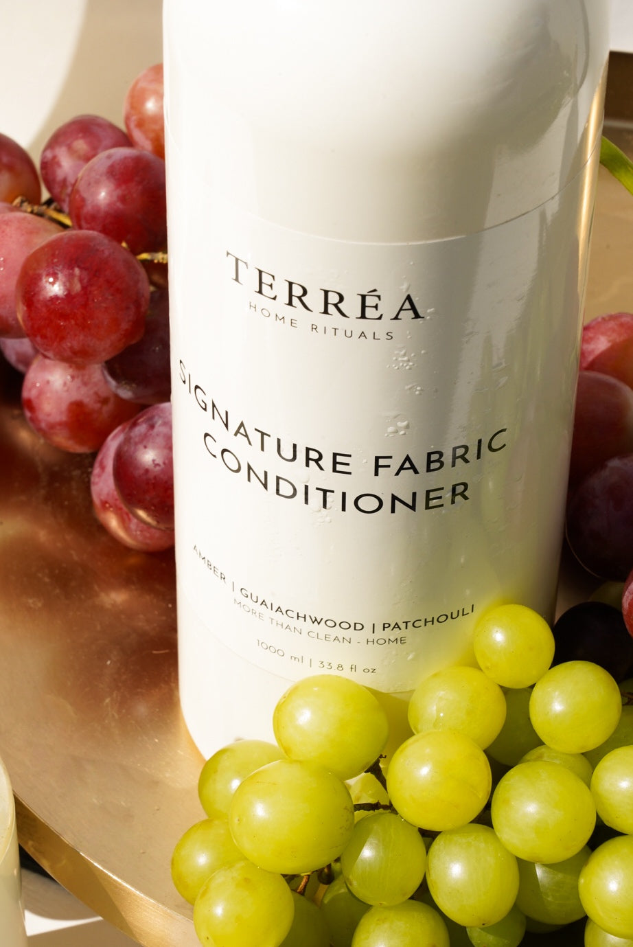 A close-up of Terréa Signature Fabric Conditioner styled with red and green grapes on a golden tray, with warm sunlight highlighting the bottle and fresh droplets.