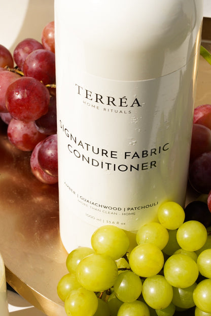 A close-up of Terréa Signature Fabric Conditioner styled with red and green grapes on a golden tray, with warm sunlight highlighting the bottle and fresh droplets.