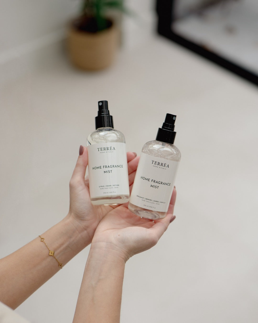Hands holding two Terréa Home Fragrance Mist bottles in Citrus, Cedar, Vetiver and Bergamot, Berries, Amber, styled against a soft neutral interior.