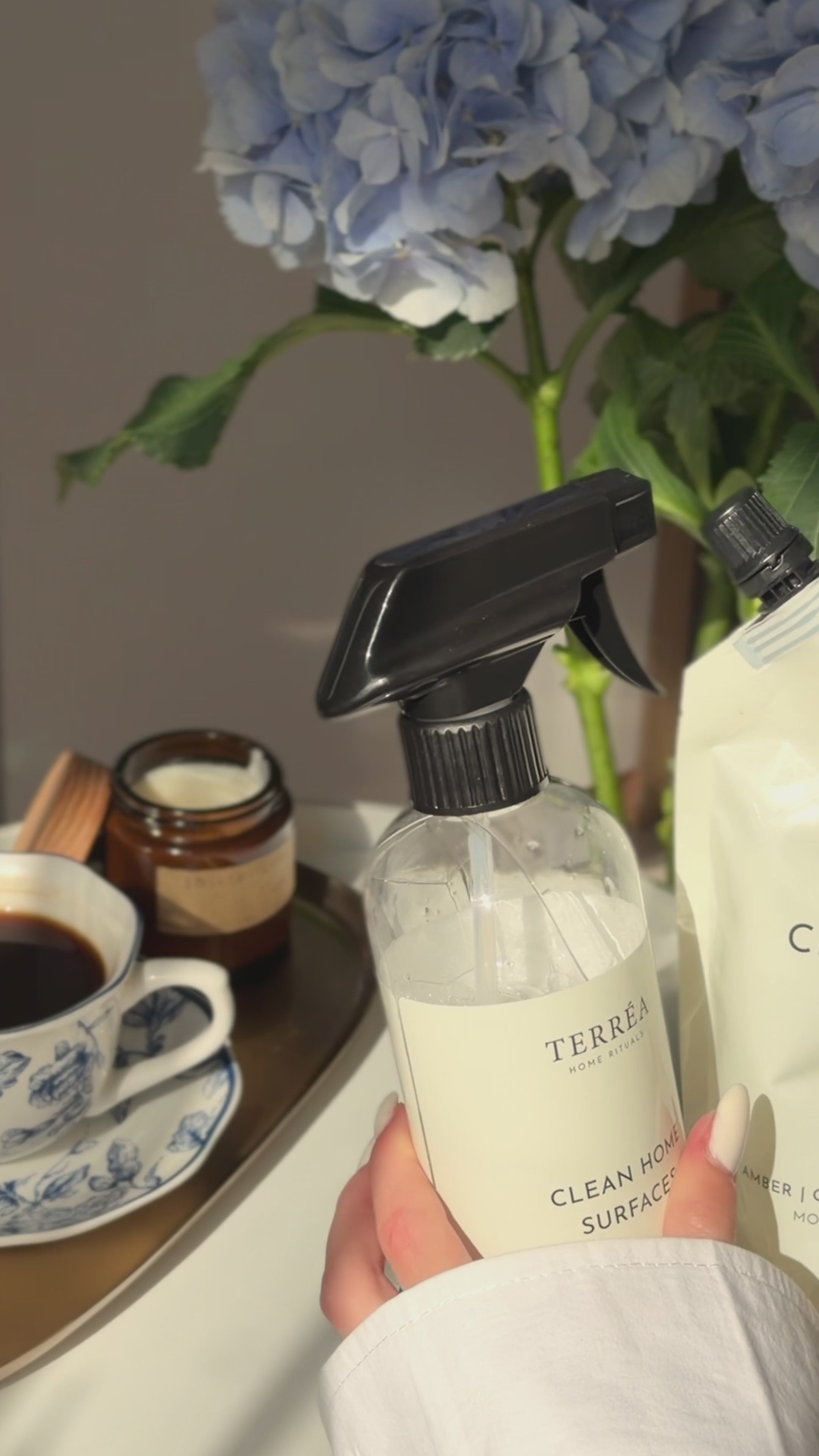 A close-up video still of Terréa Clean Home Surfaces being held beside a refill pouch, blue hydrangeas and a quiet tea tray.