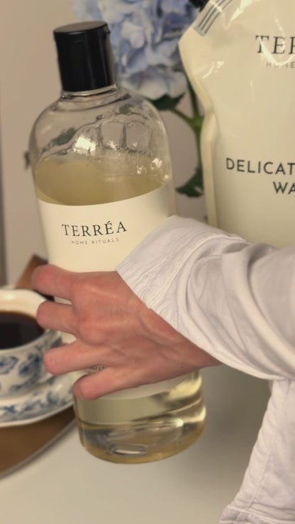A close-up video still of Terréa Delicate Fabric Wash and its refill pouch, held beside soft blue hydrangeas in a quiet laundry-care moment.
