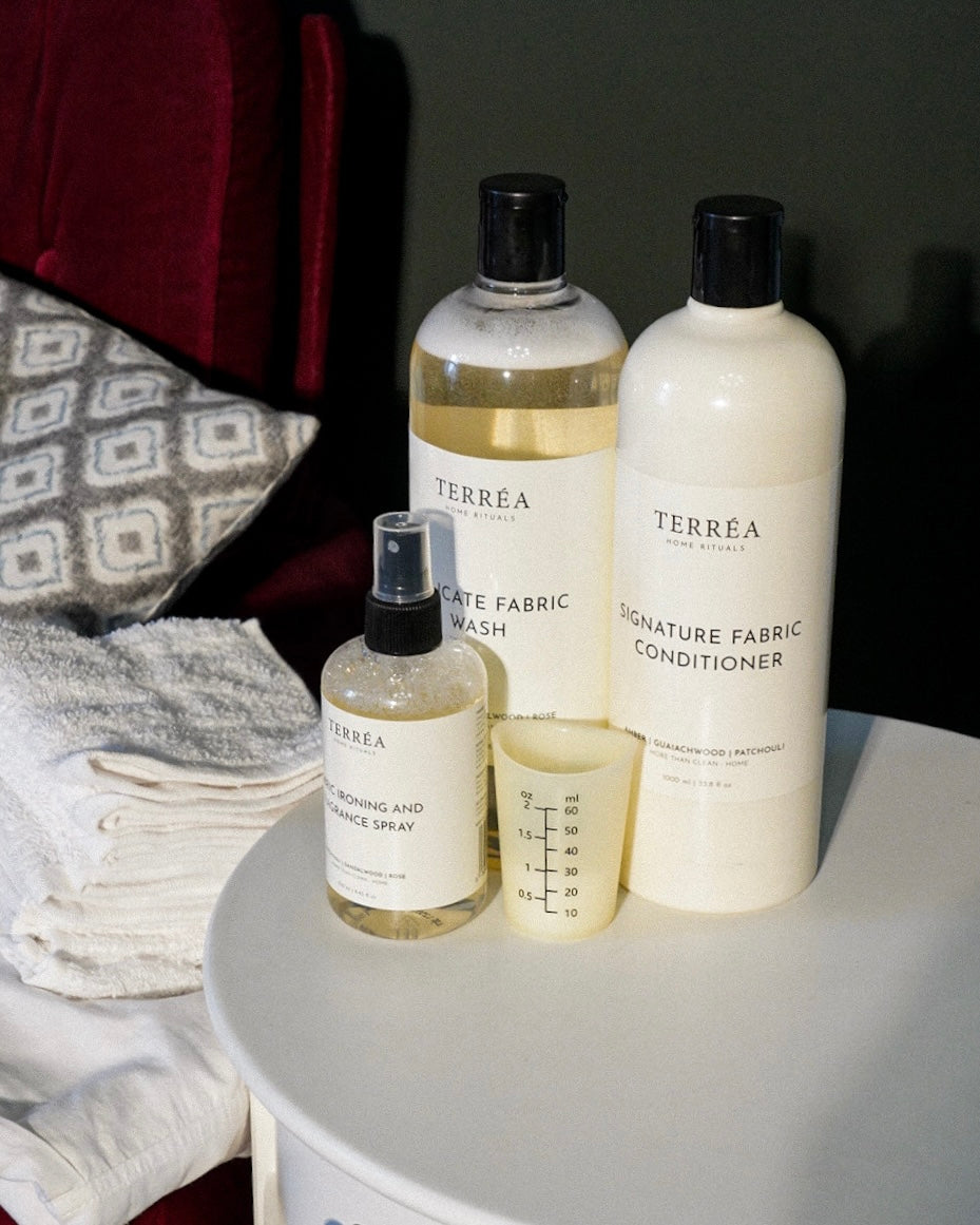 Terréa Linen Quiet laundry set on a side table in the evening: Delicate Fabric Wash, Signature Fabric Conditioner, Scented Ironing Water and the silicone measure cup, beside folded linen